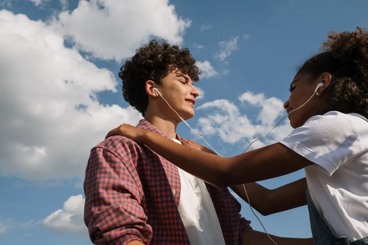 Two teens sharing earbuds and a quiet, comforting moment outdoors under a blue sky